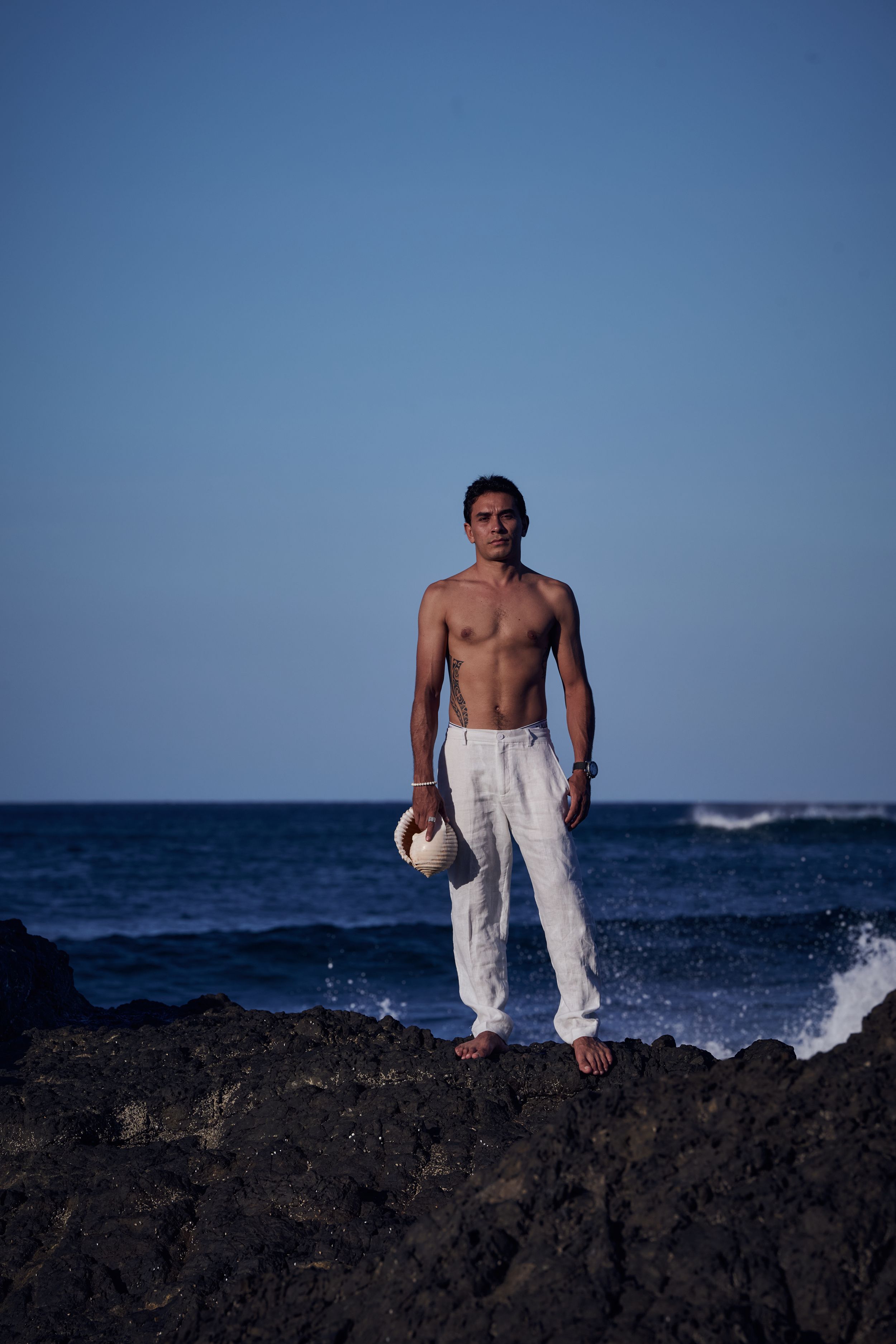 Shirtless man in white linen pants holding a hat, standing on volcanic rocks by the ocean under a clear blue sky.