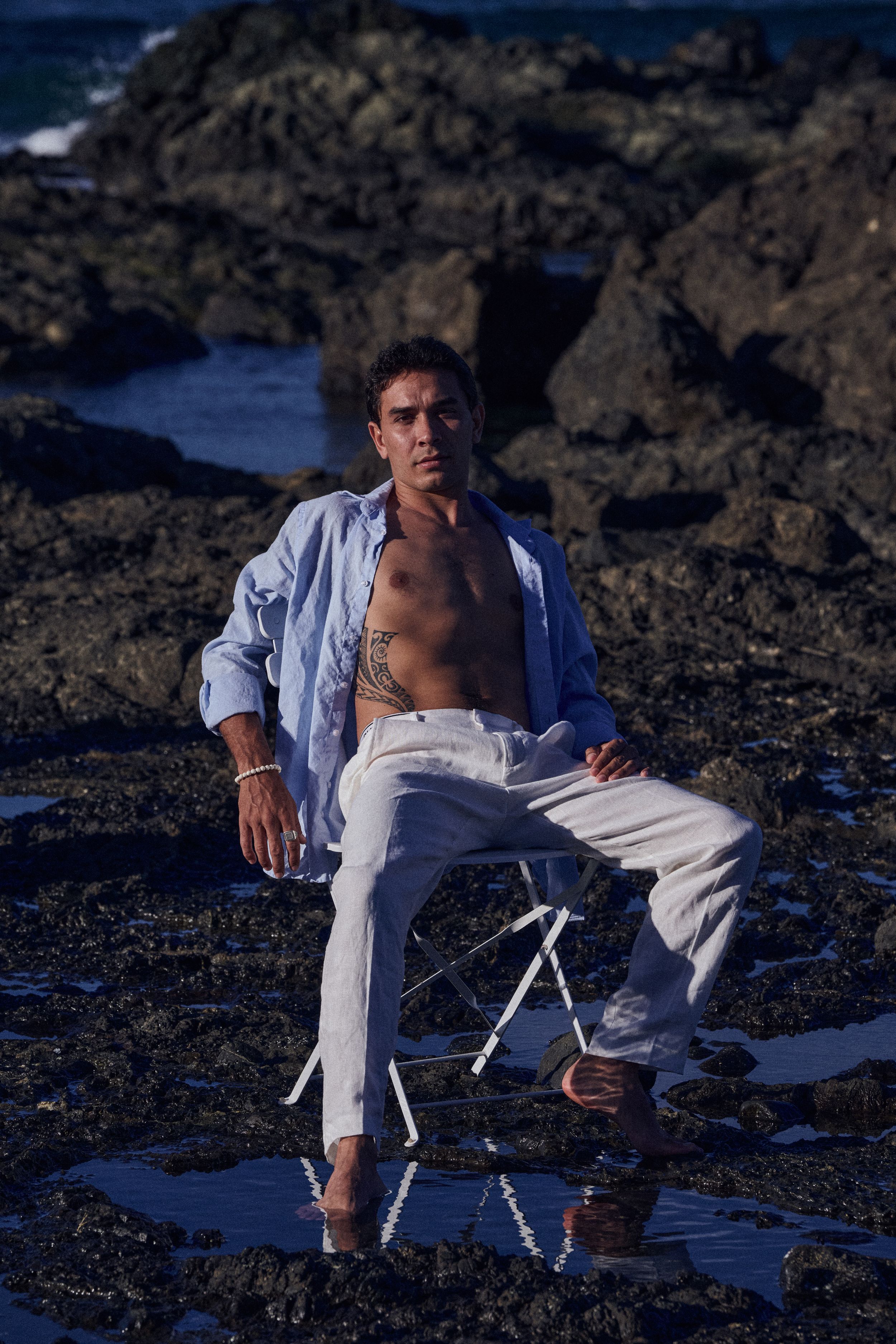 Man in open blue shirt and white pants sitting on rocks by the ocean with chest tattoo visible.