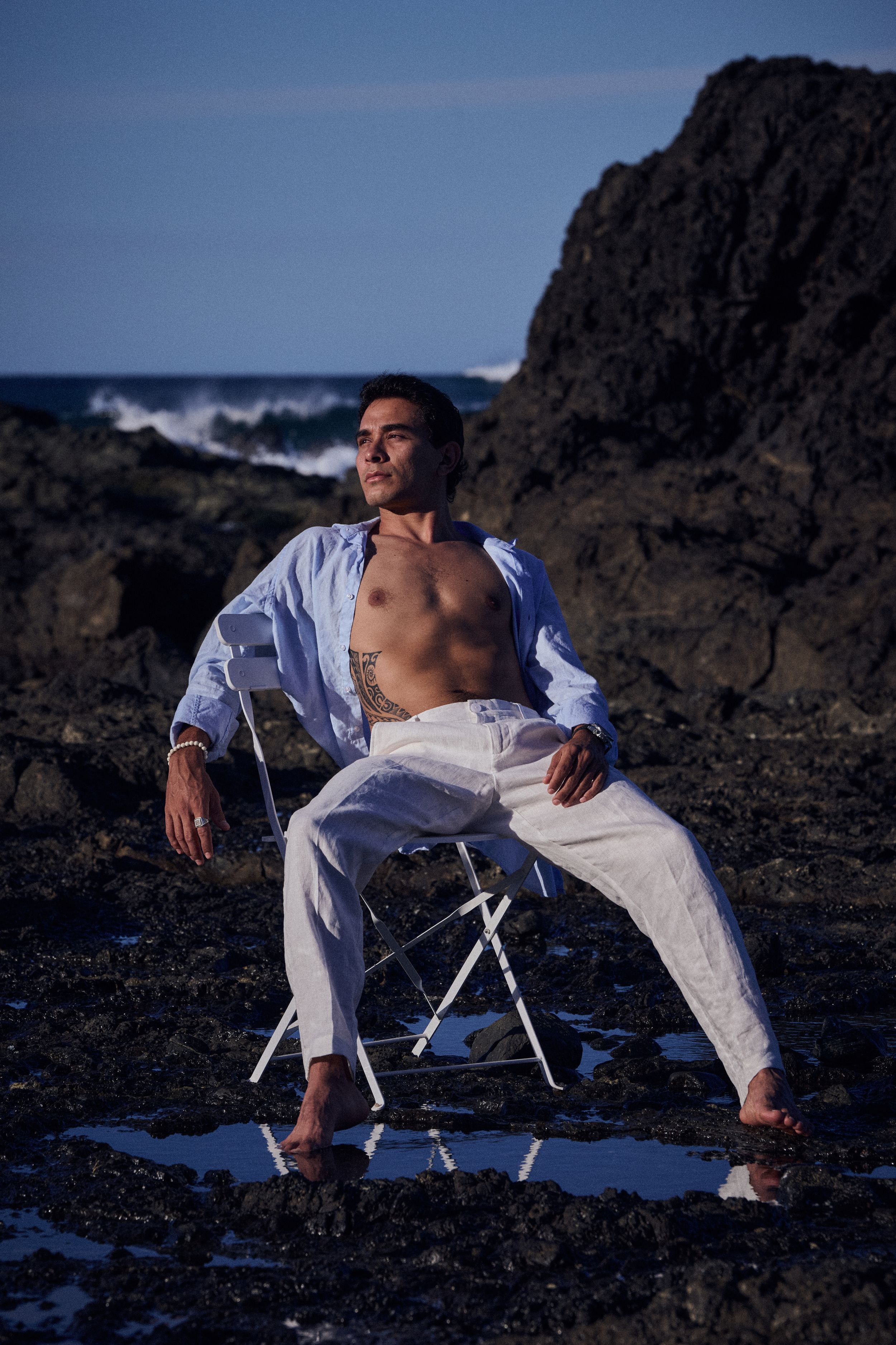 Man in open white shirt and pants sits on chair among coastal rocks with ocean waves in background.