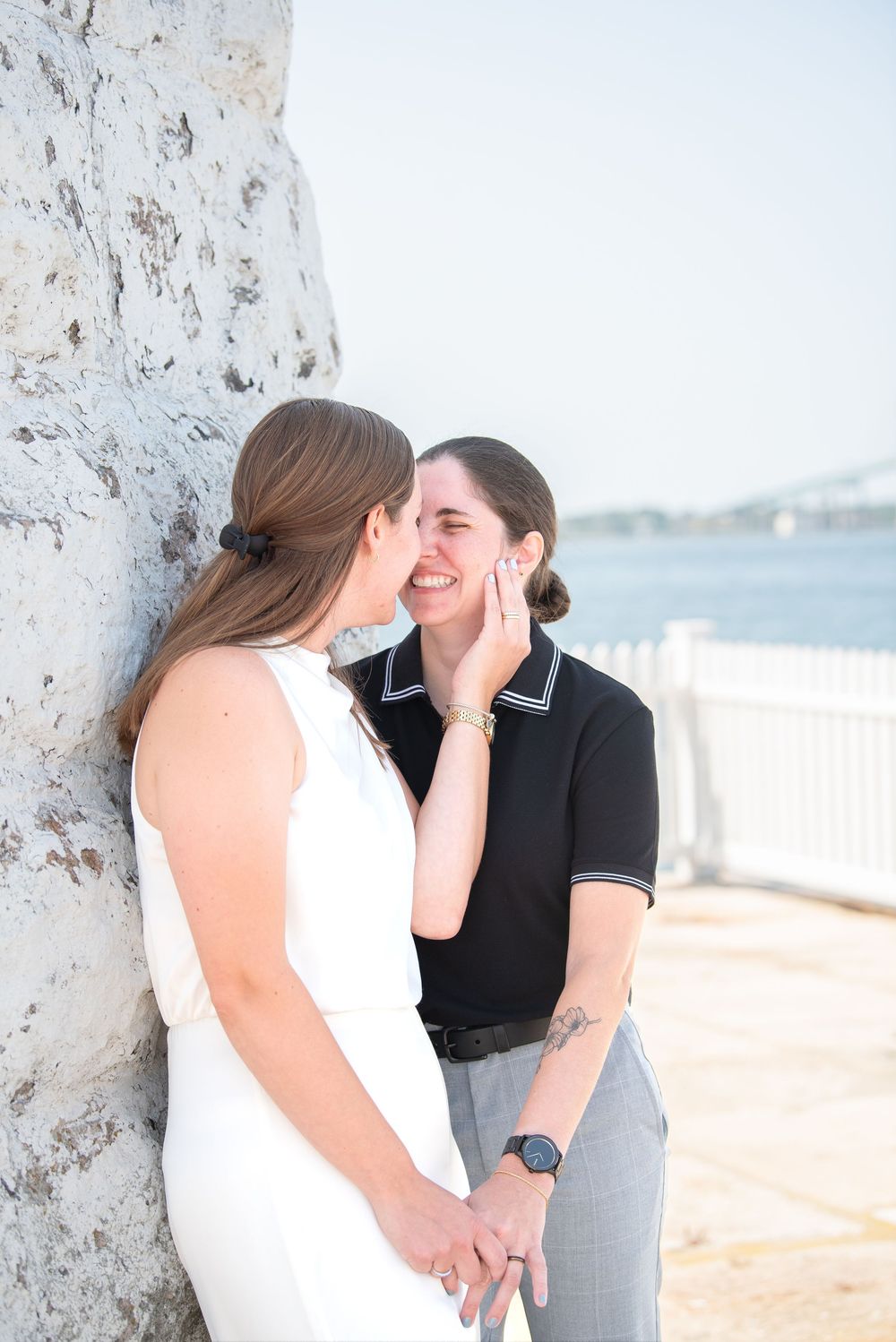 A romantic moment shared between a couple in white and black attire against a stone wall by the waterfront.