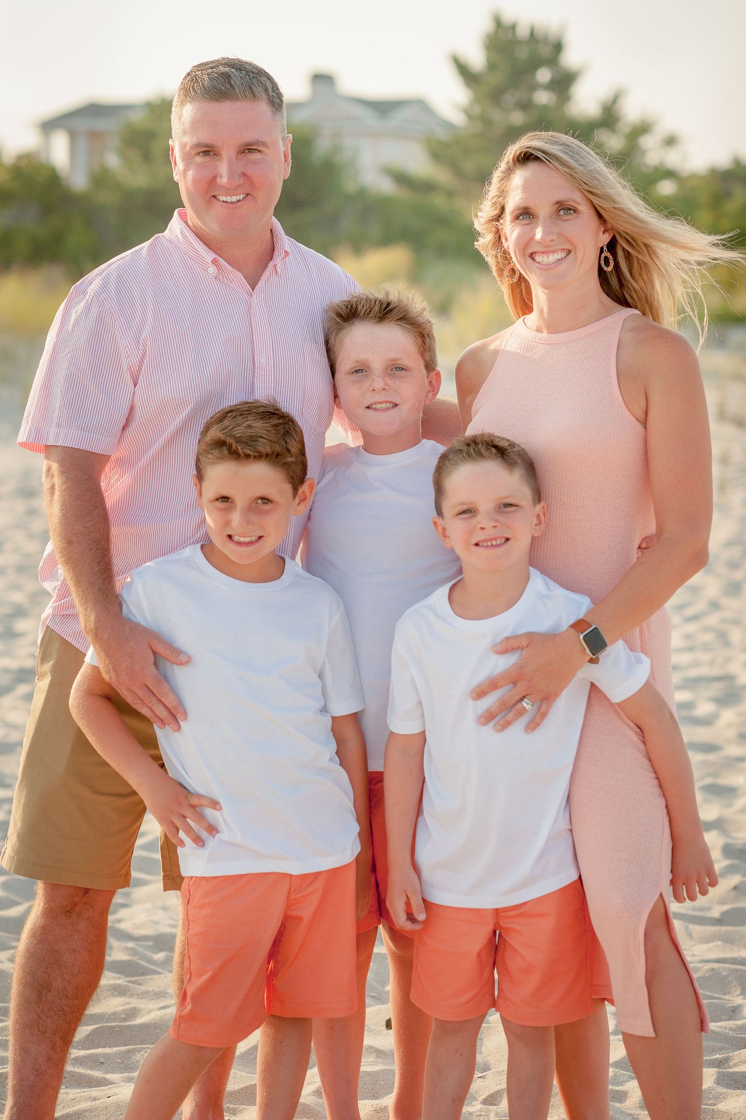 Family of five posing together on a sandy beach, parents and three boys in coordinating pink and white outfits.