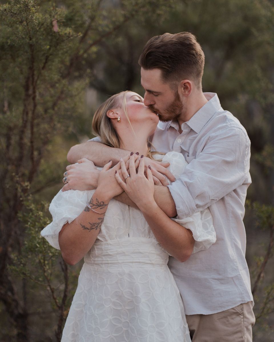 Happy couple kissing and embracing outdoors in a eucalyptus forest, woman in white dress, man in casual attire during couple photoshoot