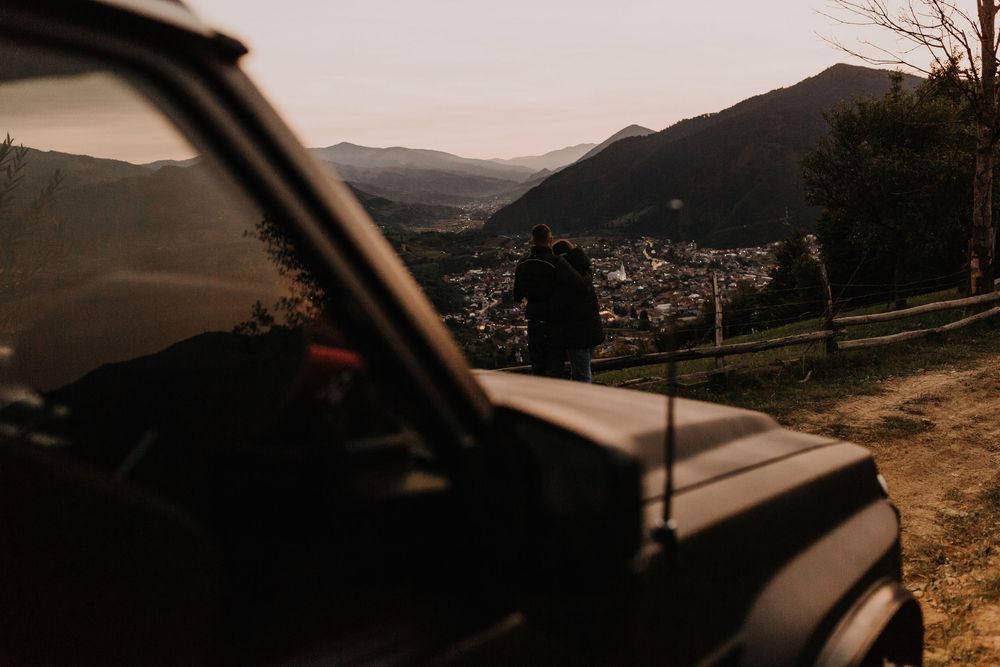 Silhouette of person standing beside black SUV overlooking mountain valley at sunset.