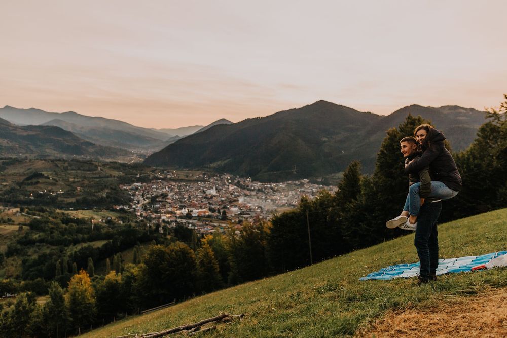 Couple enjoying piggyback ride on scenic hillside overlooking mountain town at golden sunset.