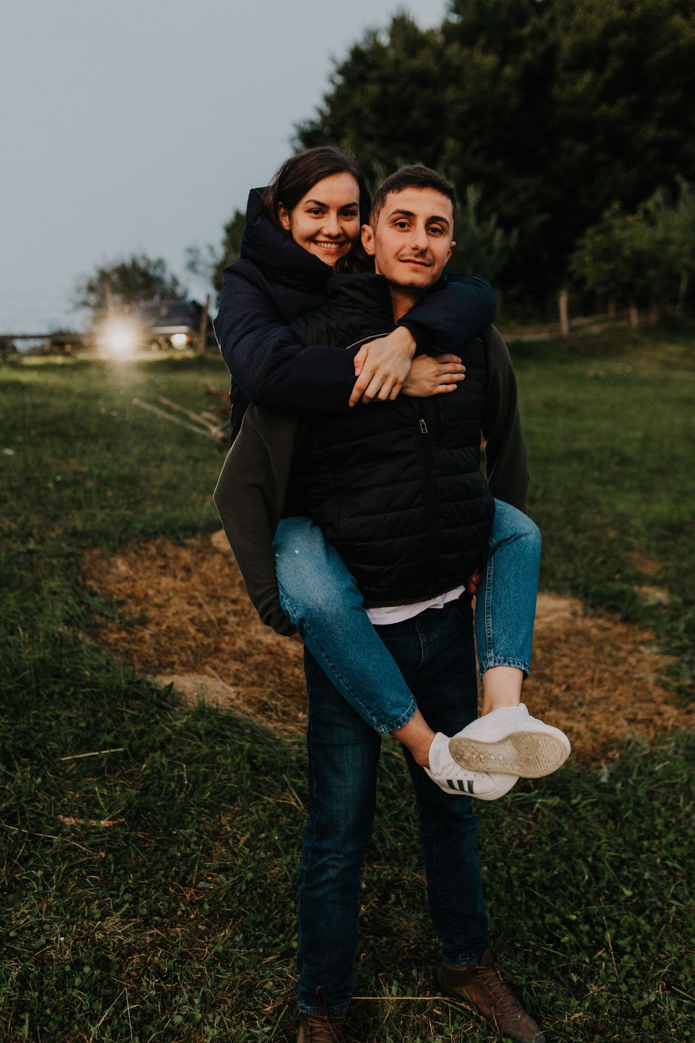Young couple smiling outdoors as man gives woman a piggyback ride in green field at dusk.