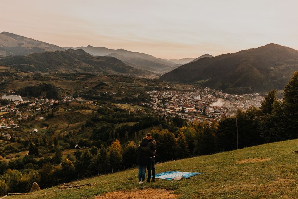 Couple standing together on hillside admiring stunning panoramic mountain town view at sunset.