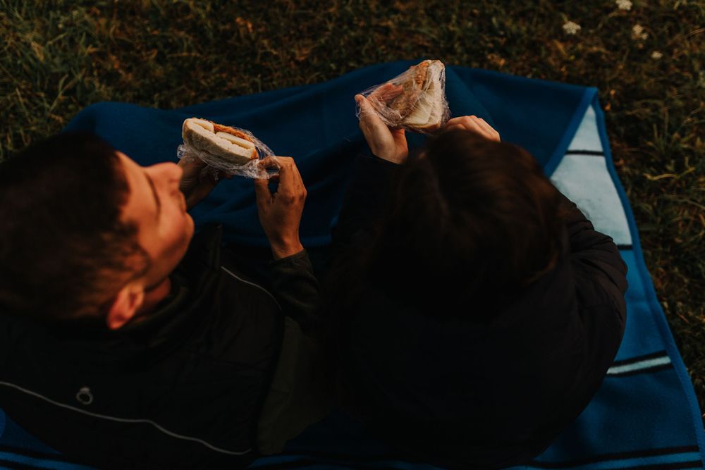 Person eating sandwiches while relaxing on a blue blanket outdoors.