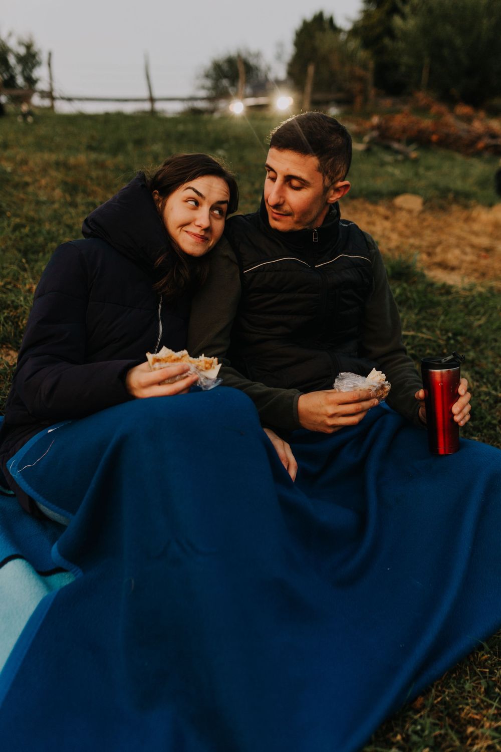 Couple sharing food and drinks wrapped in a blue blanket at twilight.