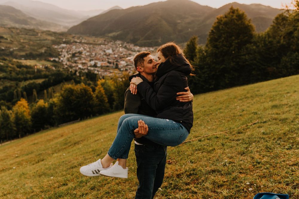 Man lifting and kissing woman on a green hillside with mountain backdrop.