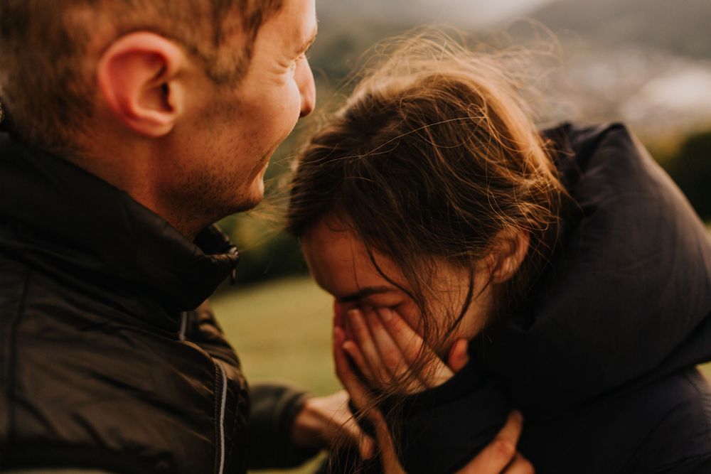 An emotional young couple shares an intimate moment outdoors, with the woman hiding her face.