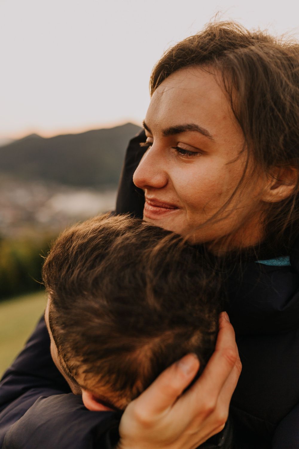 A smiling young woman tenderly embraces her partner outdoors with scenic mountains in the background.