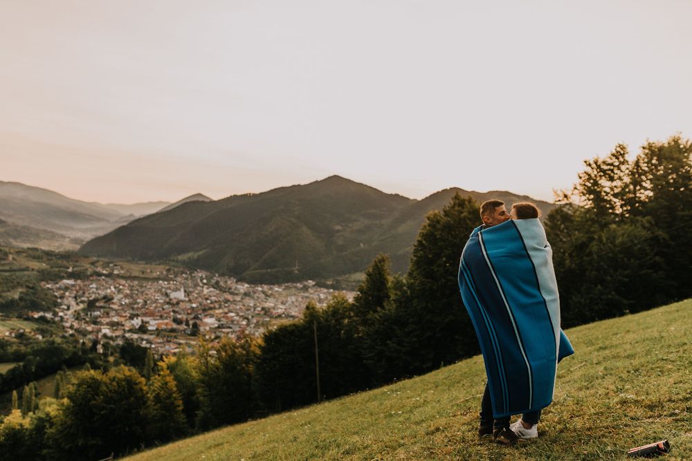 A romantic couple wrapped in a blue blanket stands on a hilltop overlooking a mountain valley town.