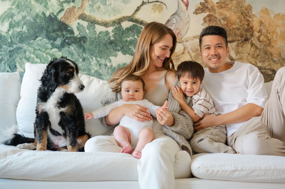 Happy family with two babies and a dog posing on a white couch in front of a floral wallpaper backdrop.