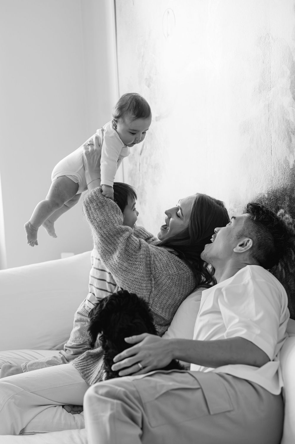 Black and white family photo of parents lifting baby on couch with toddler and dog.