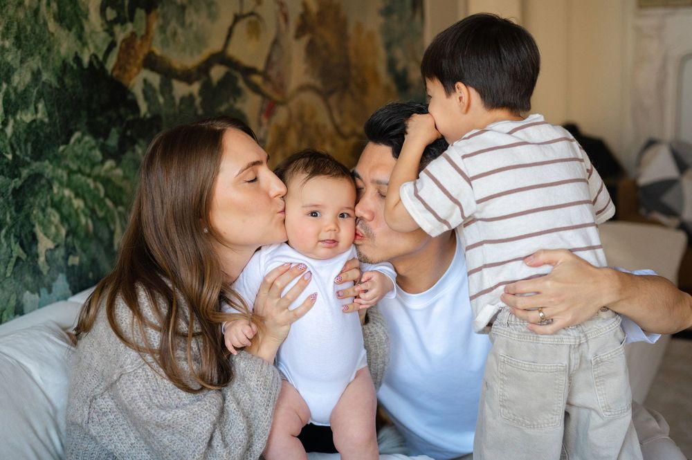 Parents kissing their newborn baby while older child hugs dad in a cozy home.