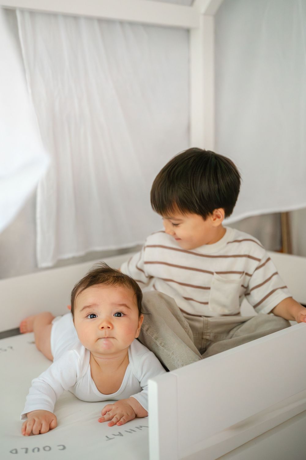 Baby in white onesie lying in crib with older toddler brother looking on.