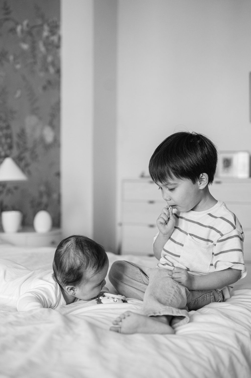 Black and white photo of a young boy sitting on a bed looking at his newborn sibling.