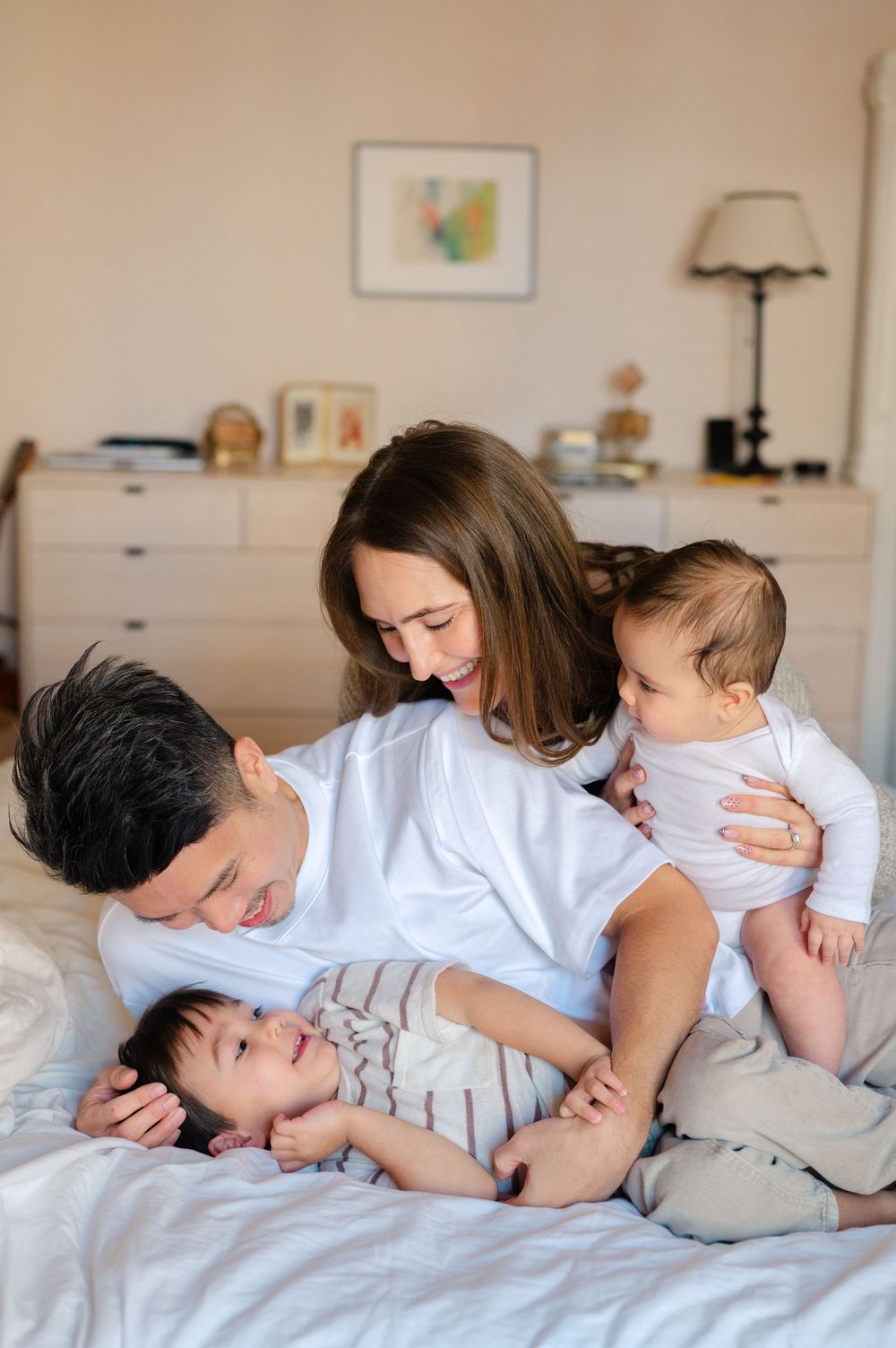 Family of four with two babies enjoying a cozy moment together on a white bed in a bright bedroom.