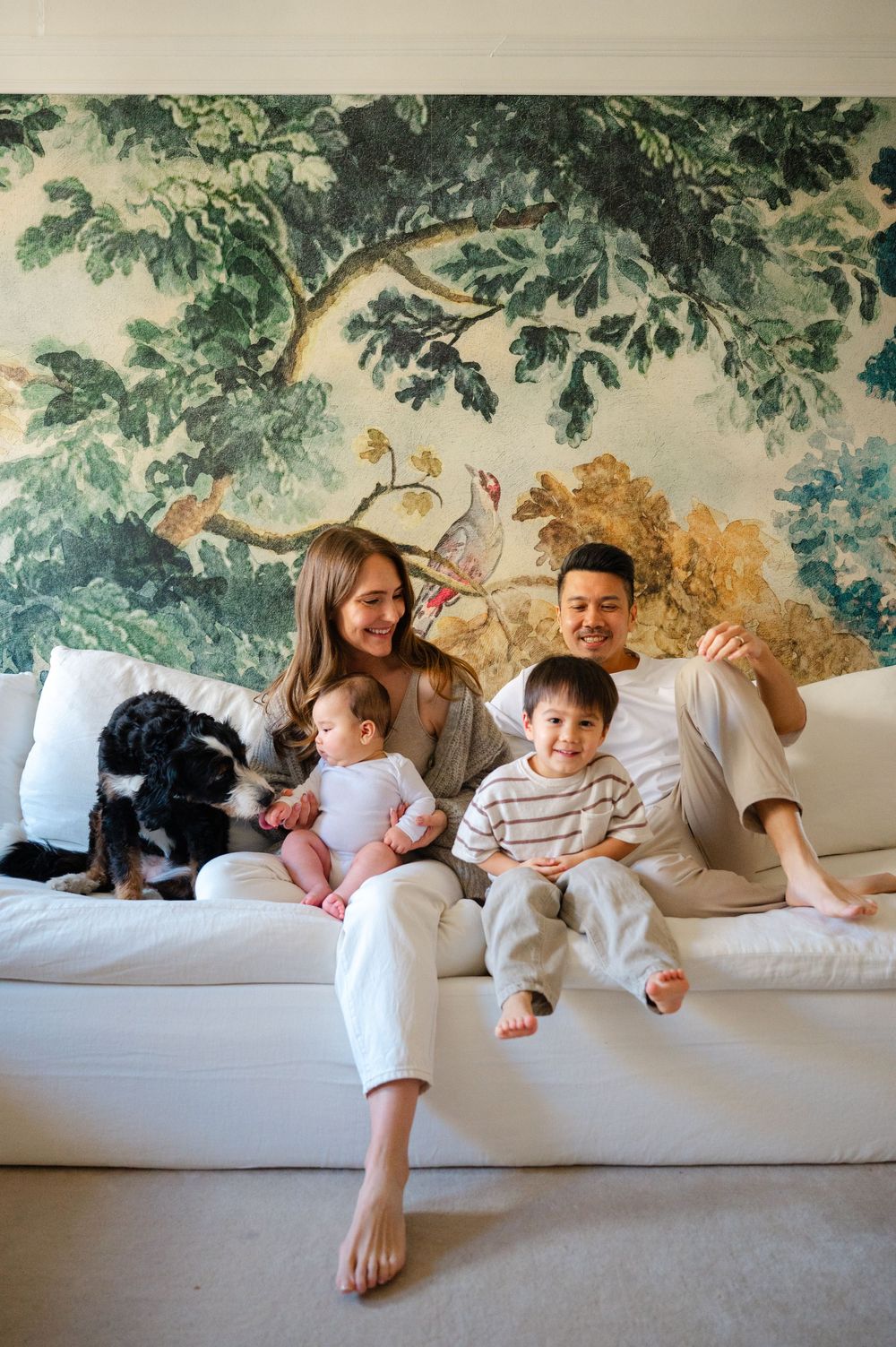 Happy family with two young children and a dog sitting on a white sofa in front of a lush green botanical wallpaper mural.