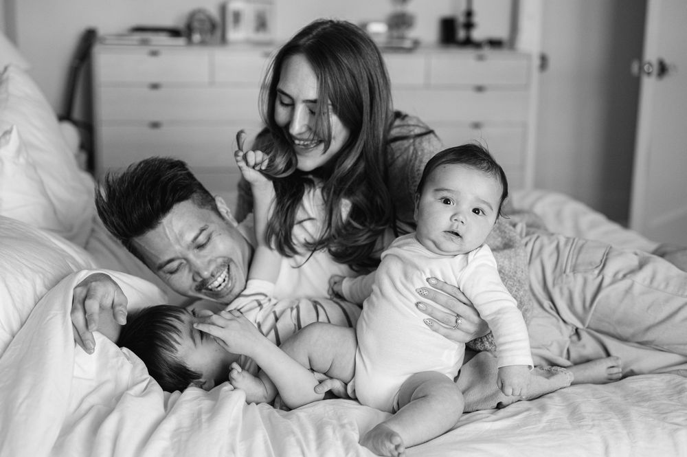Black and white photo of happy parents bonding with their two babies on a cozy white bed.