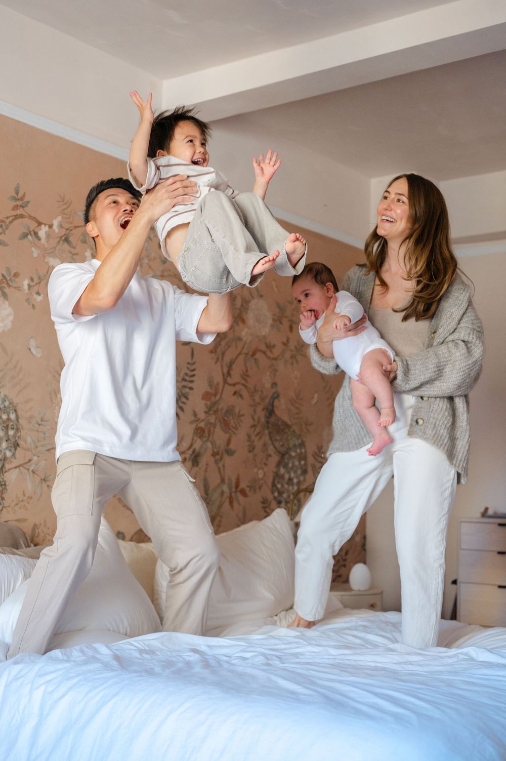 Joyful family jumping on bed, dad lifting toddler while mom holds newborn baby.