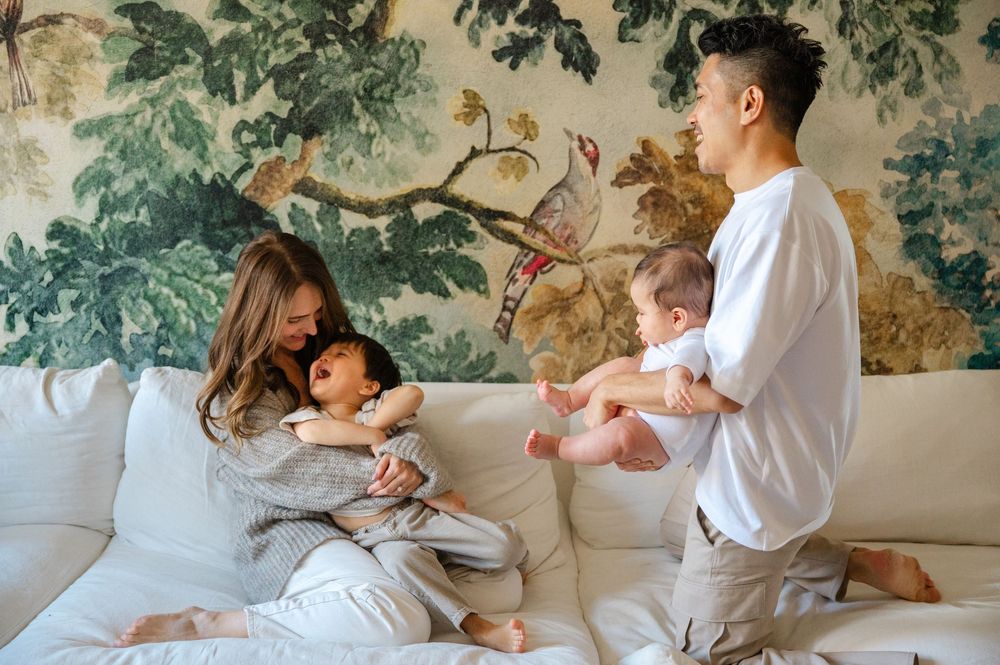 Young parents sitting on a white sofa holding their two babies in front of a floral wallpaper.