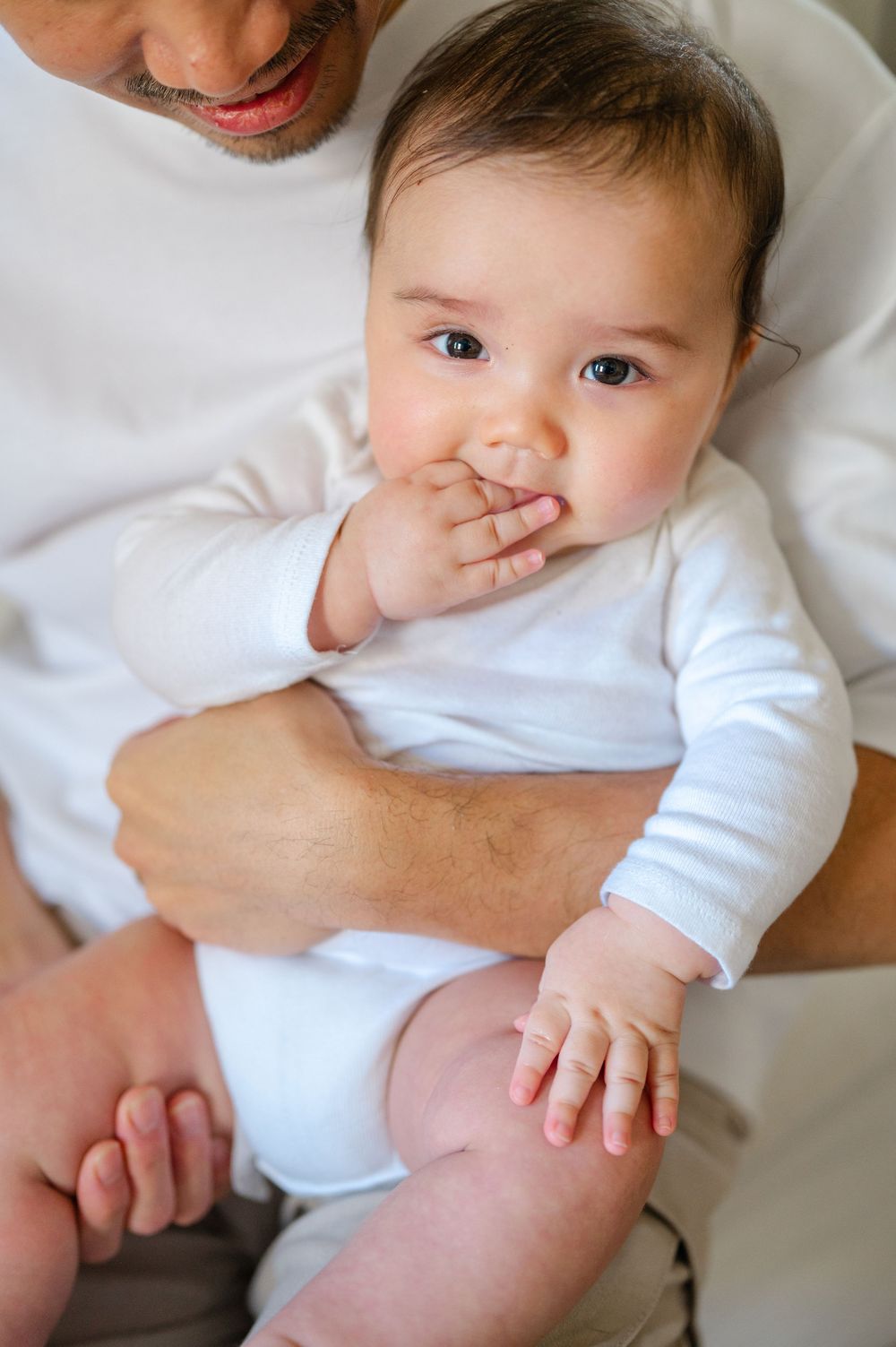 Adorable baby in white onesie chewing fingers while being held by father on a bed.