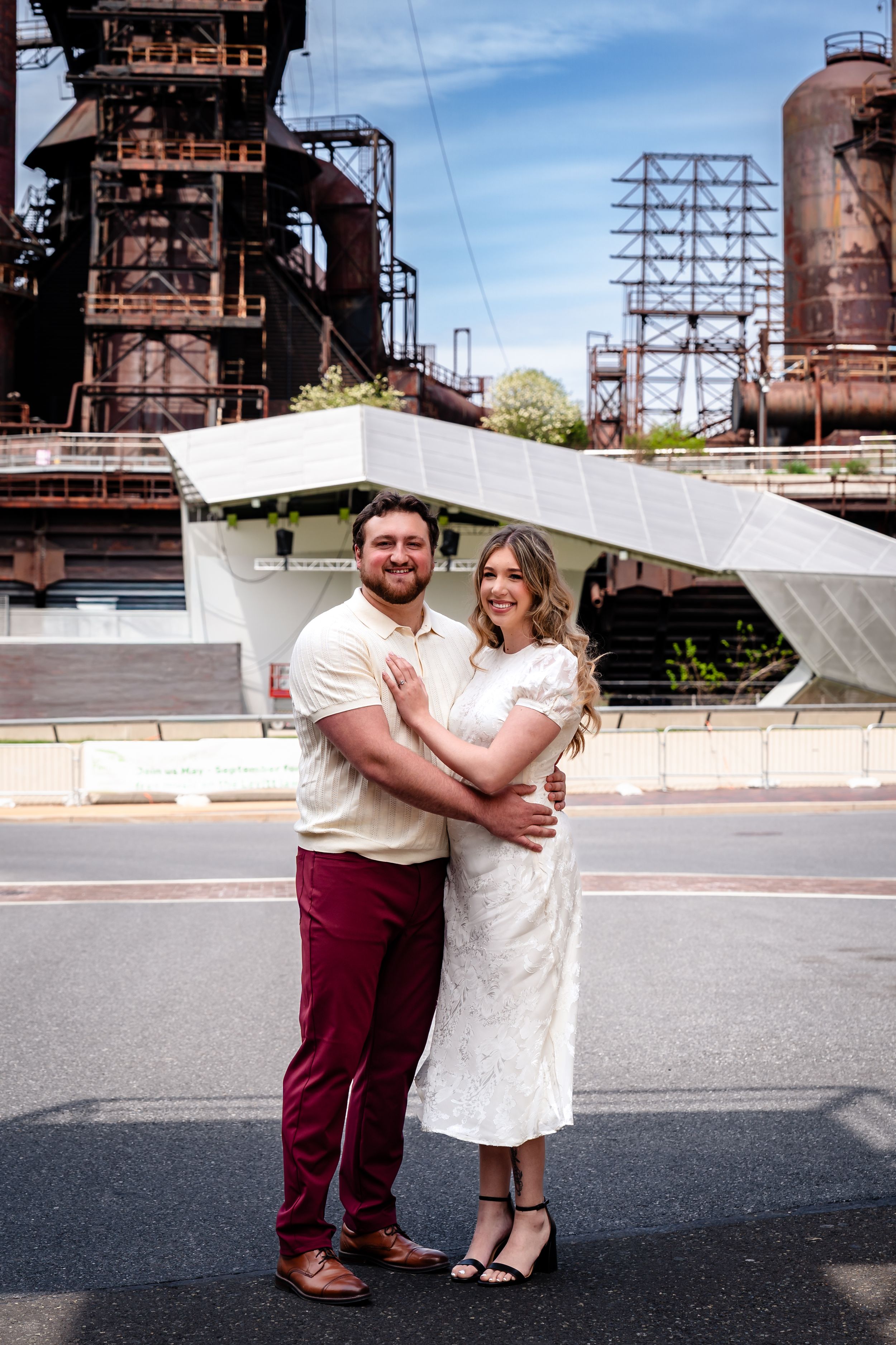 Couple embracing in white dress and burgundy pants posing before industrial steel mill backdrop on sunny day.