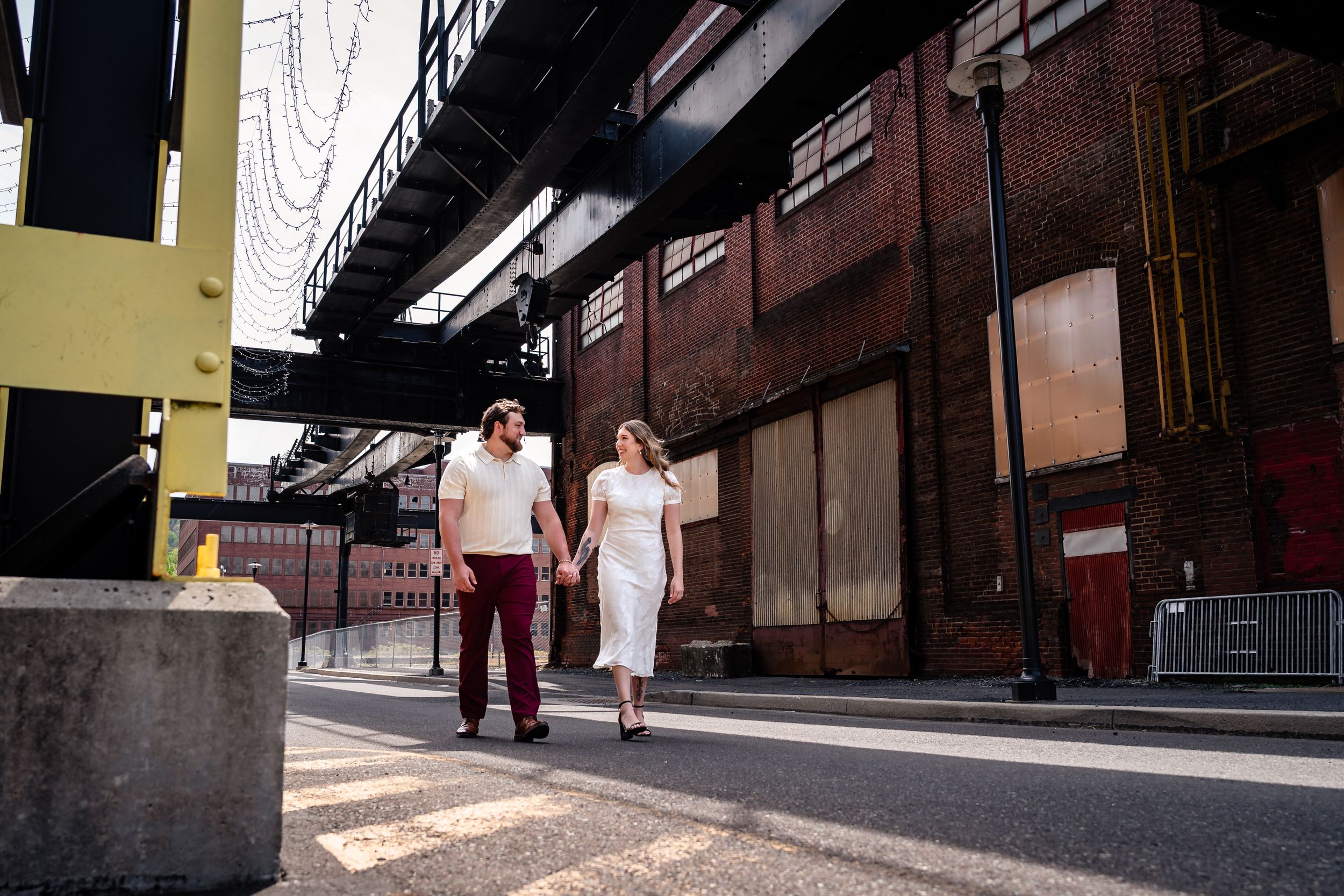 Couple walking hand in hand through an urban industrial area with brick buildings and overhead structure.