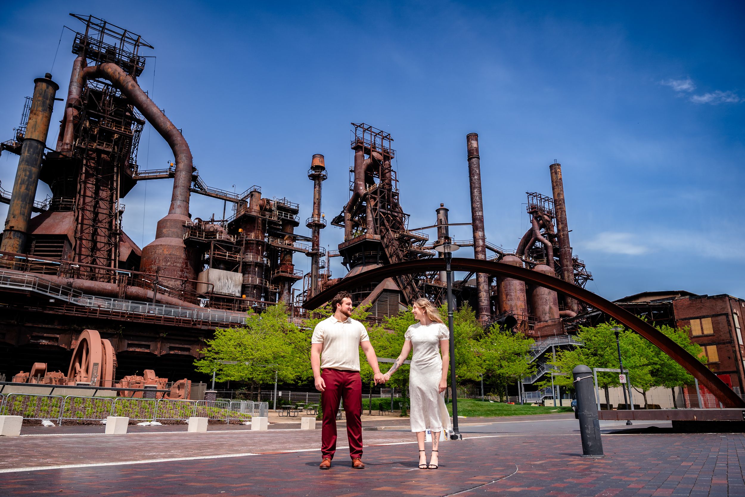 Couple holding hands in front of historic Bethlehem Steel blast furnaces under a bright blue sky.