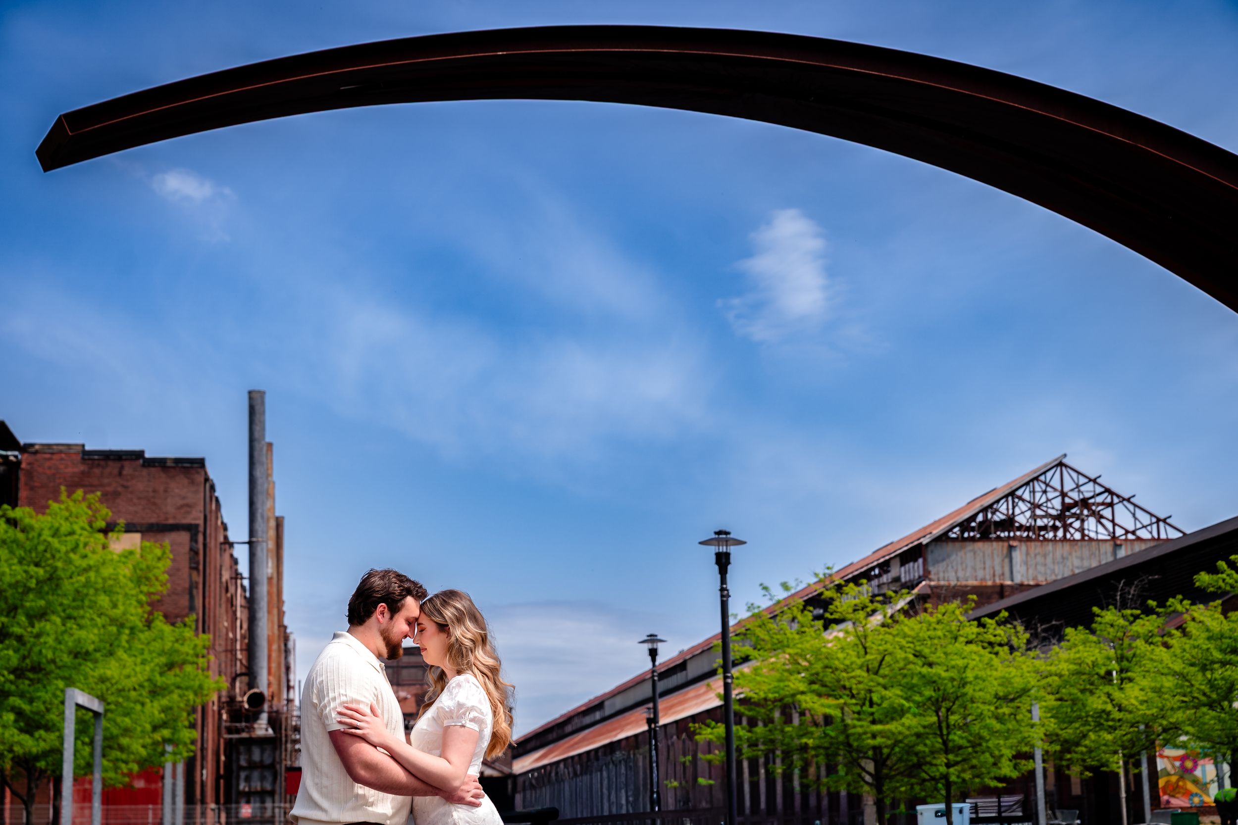 Couple embracing under an arch with blue sky, green trees, and industrial buildings in the background.