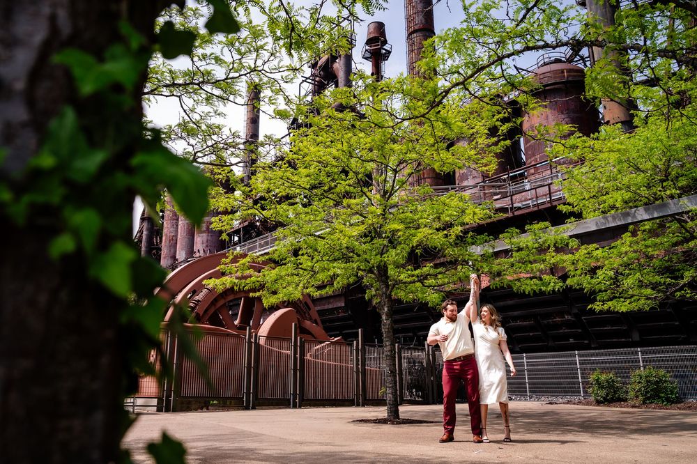 Couple walking near rustic industrial structure surrounded by lush green trees on a sunny day.