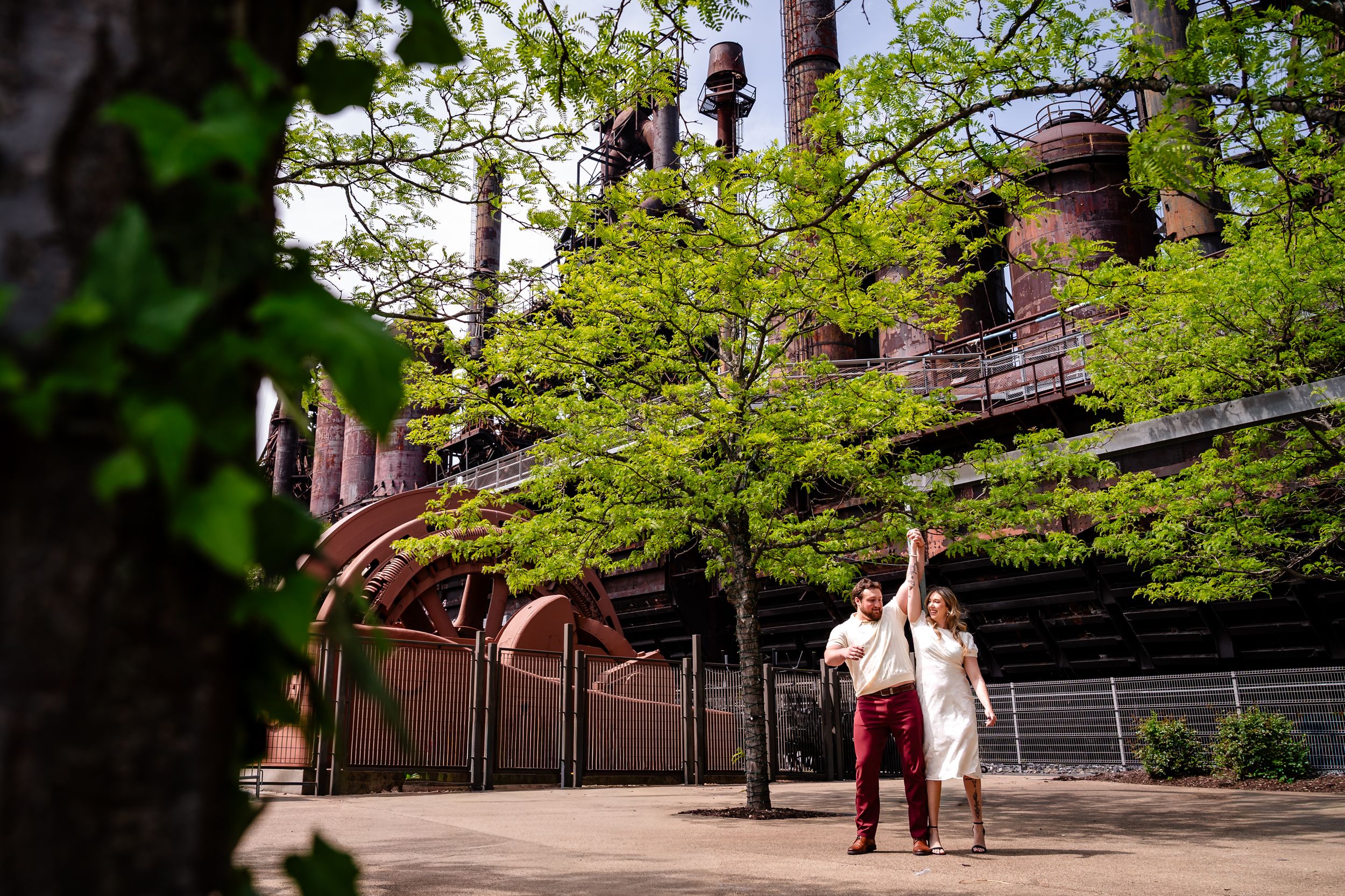 Couple walking near rustic industrial structure surrounded by lush green trees on a sunny day.