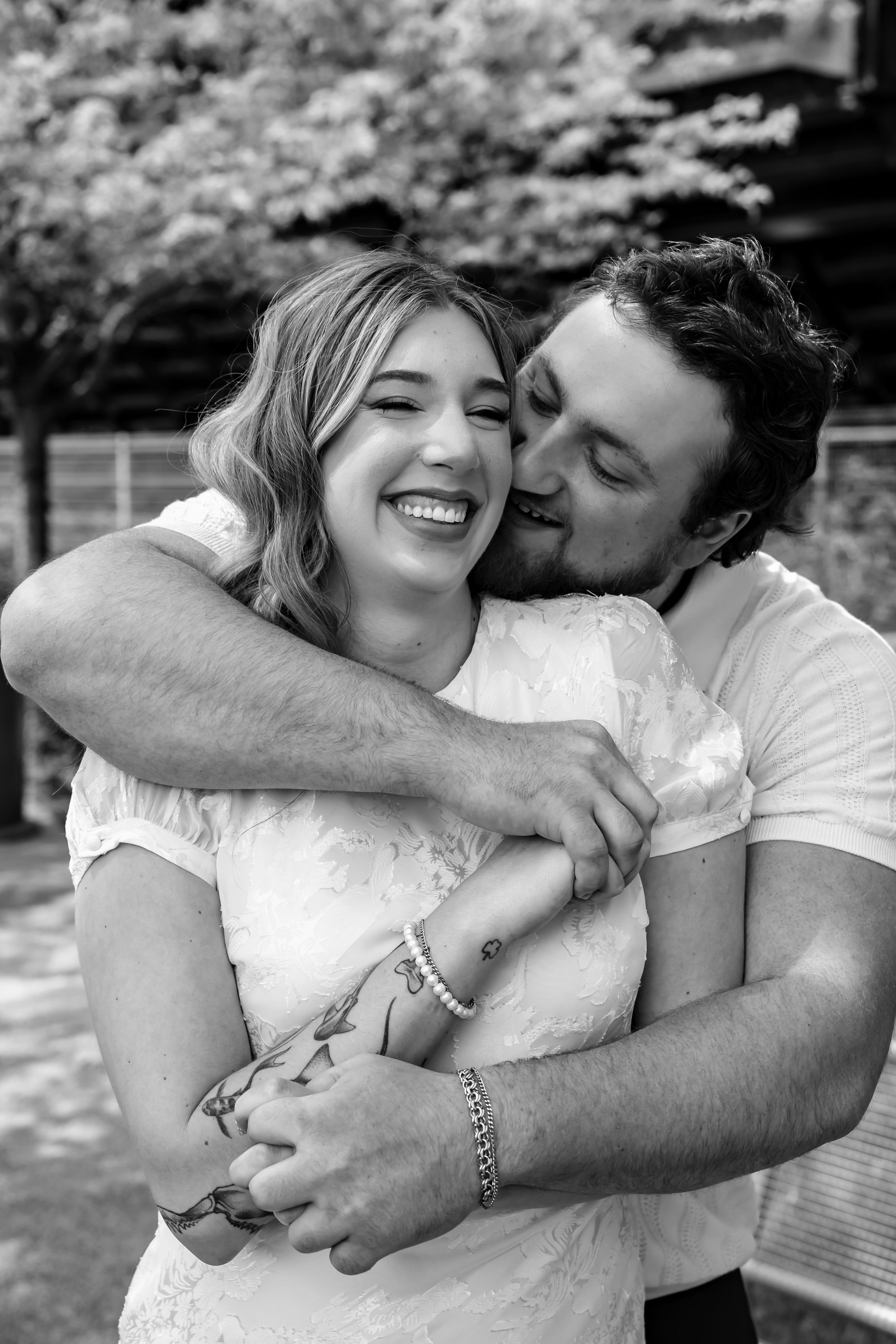 Black and white photo of a smiling couple embracing outdoors, woman laughing joyfully as man kisses her cheek.