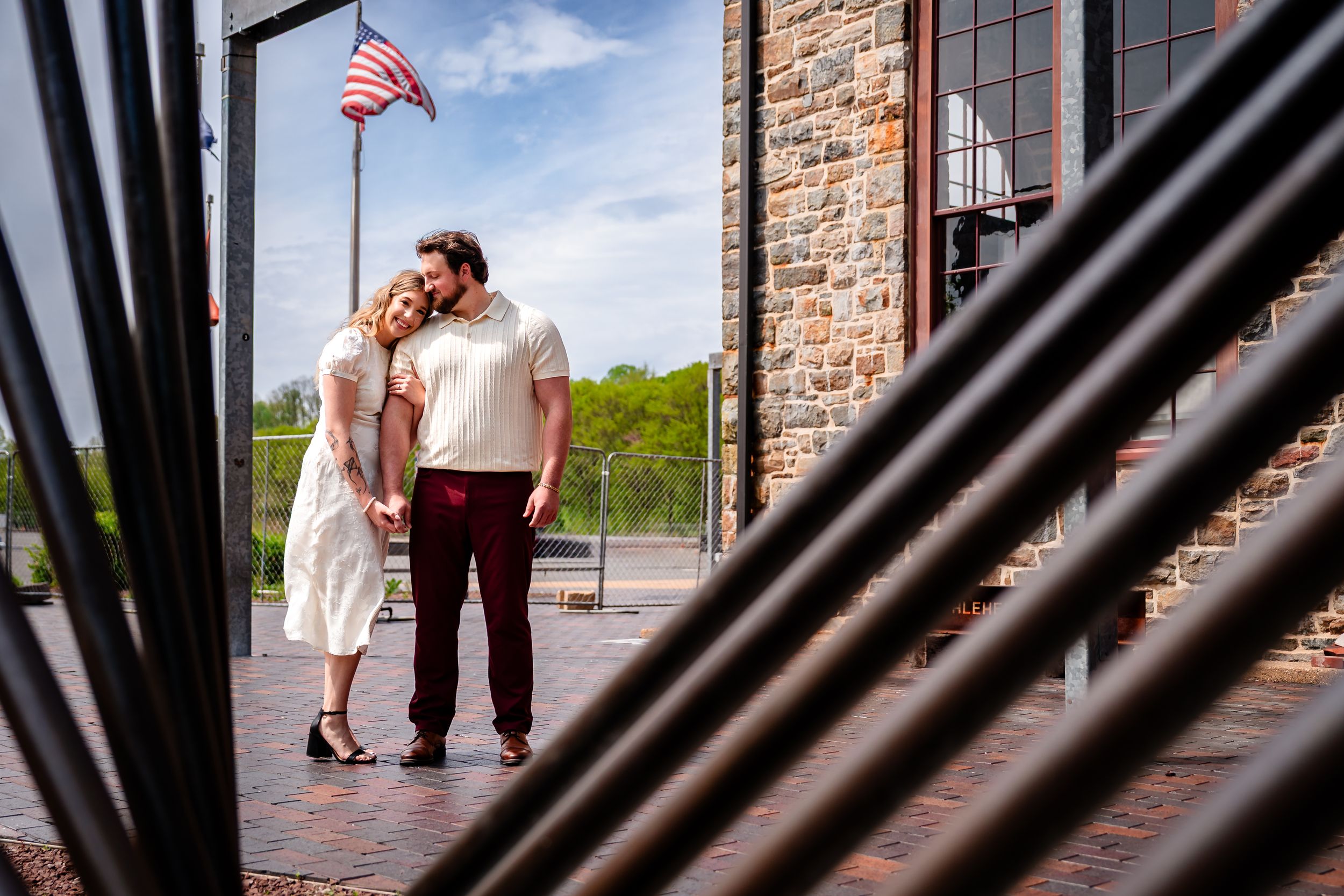 Couple sharing a romantic moment on a brick patio near a stone building with an American flag on a sunny day.