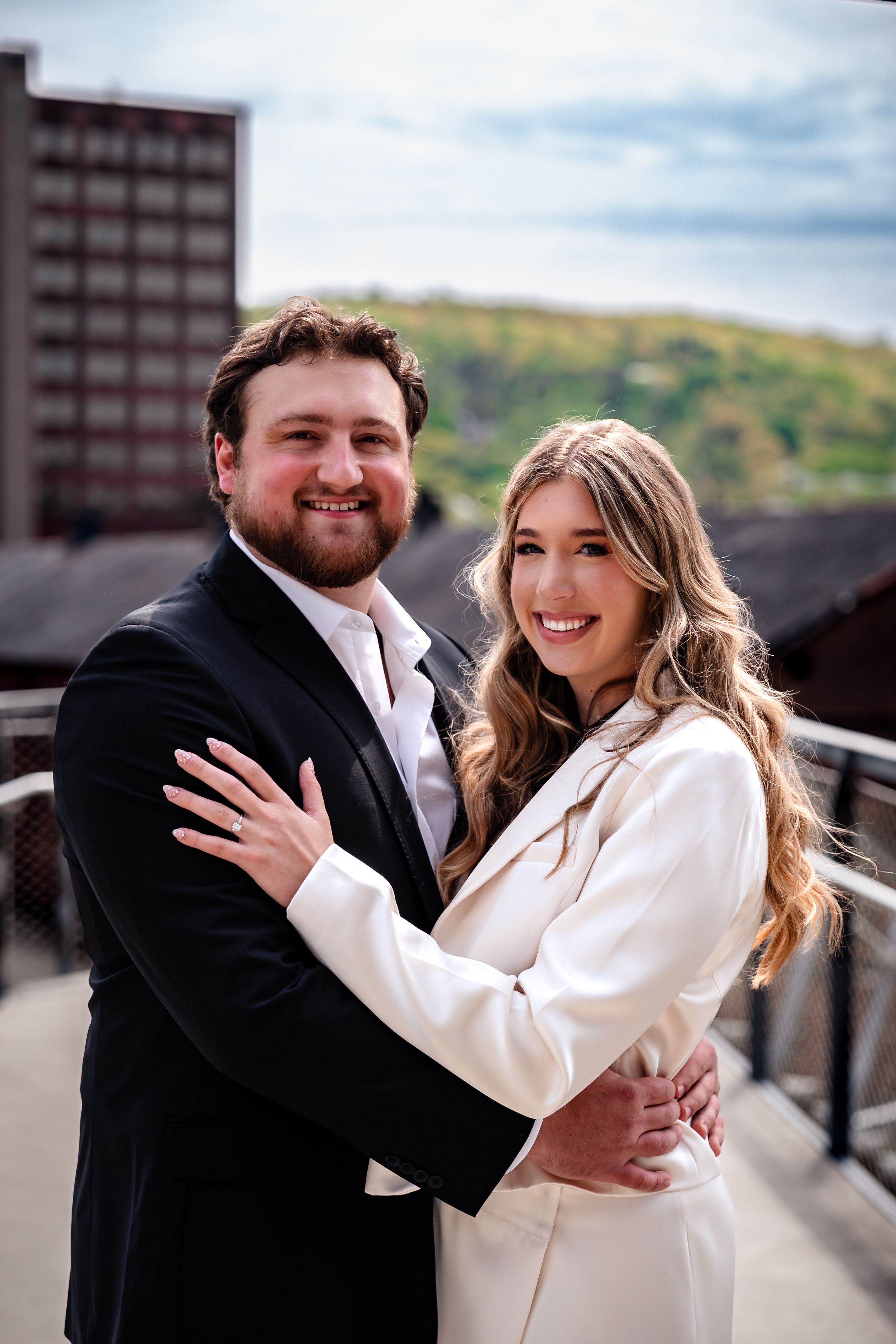 Smiling couple embracing on a bridge with city skyline and hills in the background.