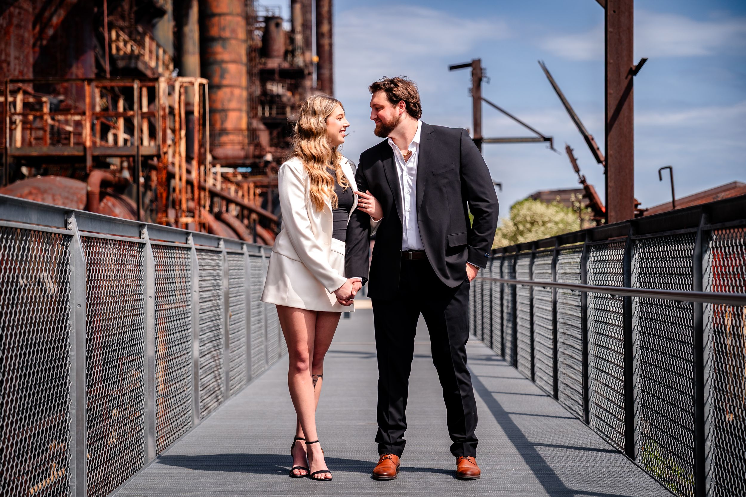 Couple holding hands on a bridge walkway near rustic industrial structures under a blue sky.