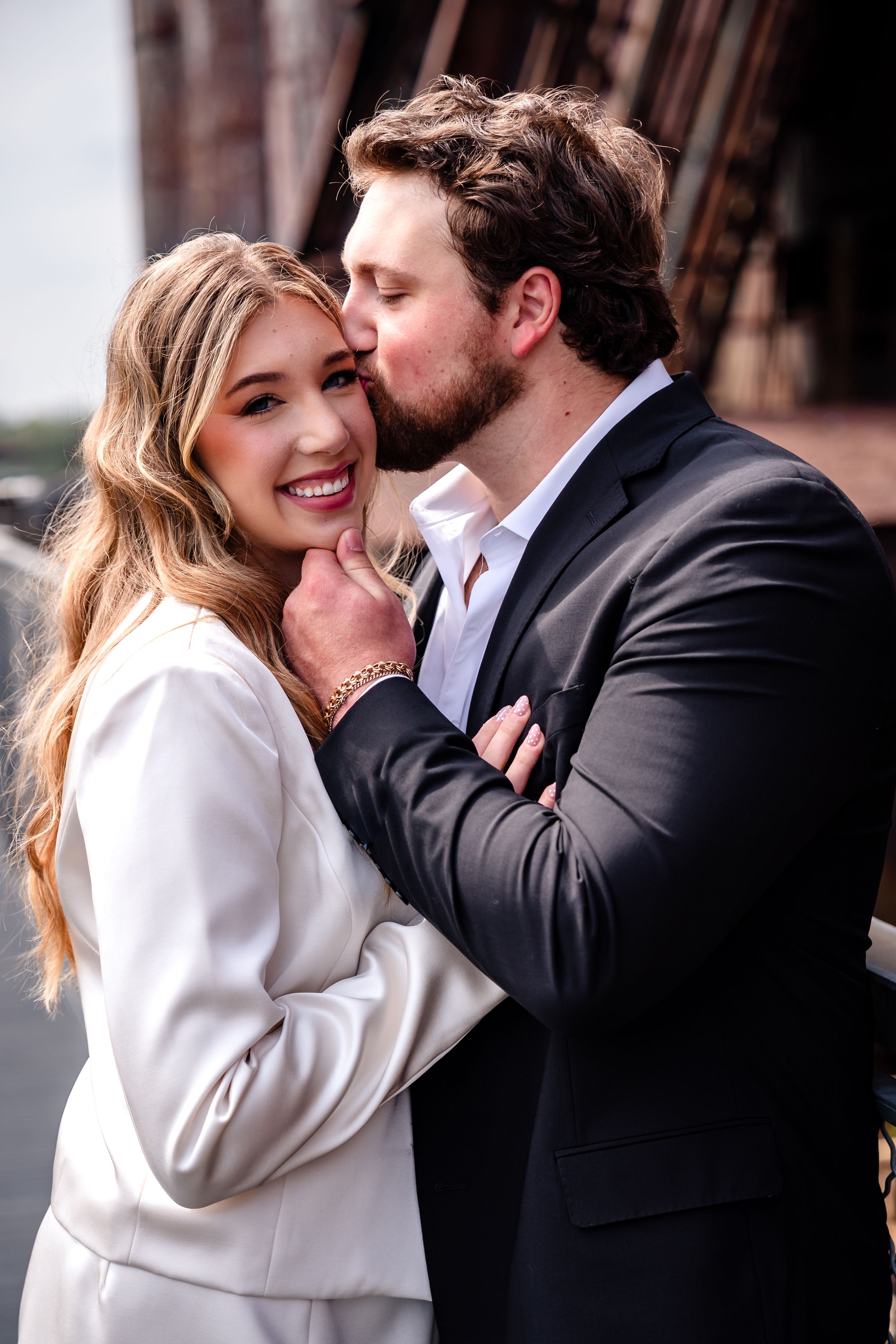 A bearded man in a dark suit kisses a smiling blonde woman in a white blazer during a romantic couples photo session.