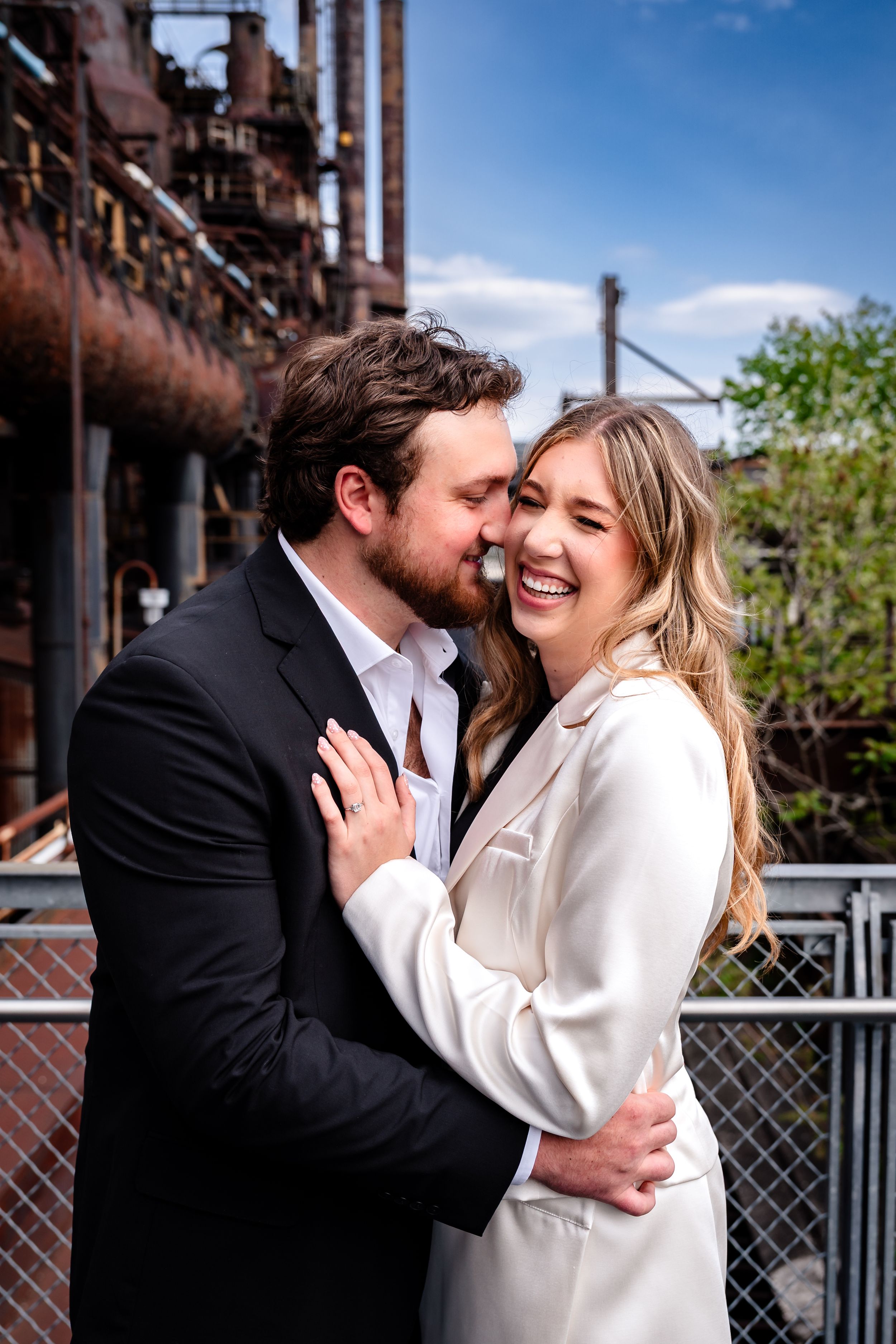 Couple embracing and laughing in front of rustic industrial backdrop, woman in white blazer, man in dark suit.