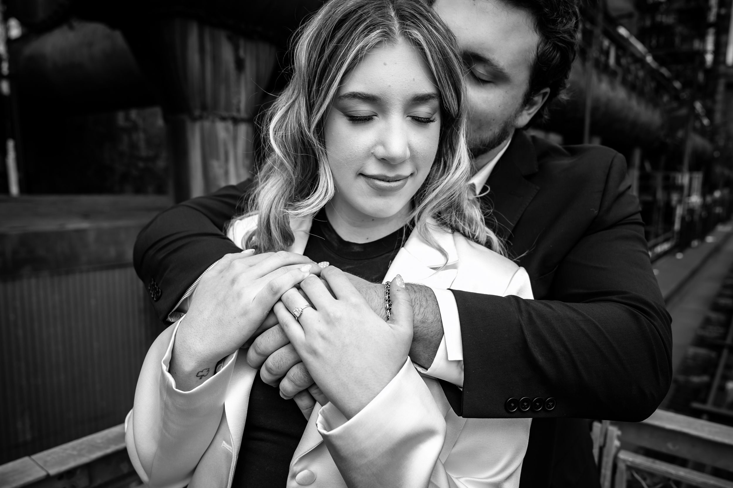 Romantic black and white photo of a man embracing a woman from behind at an industrial setting.