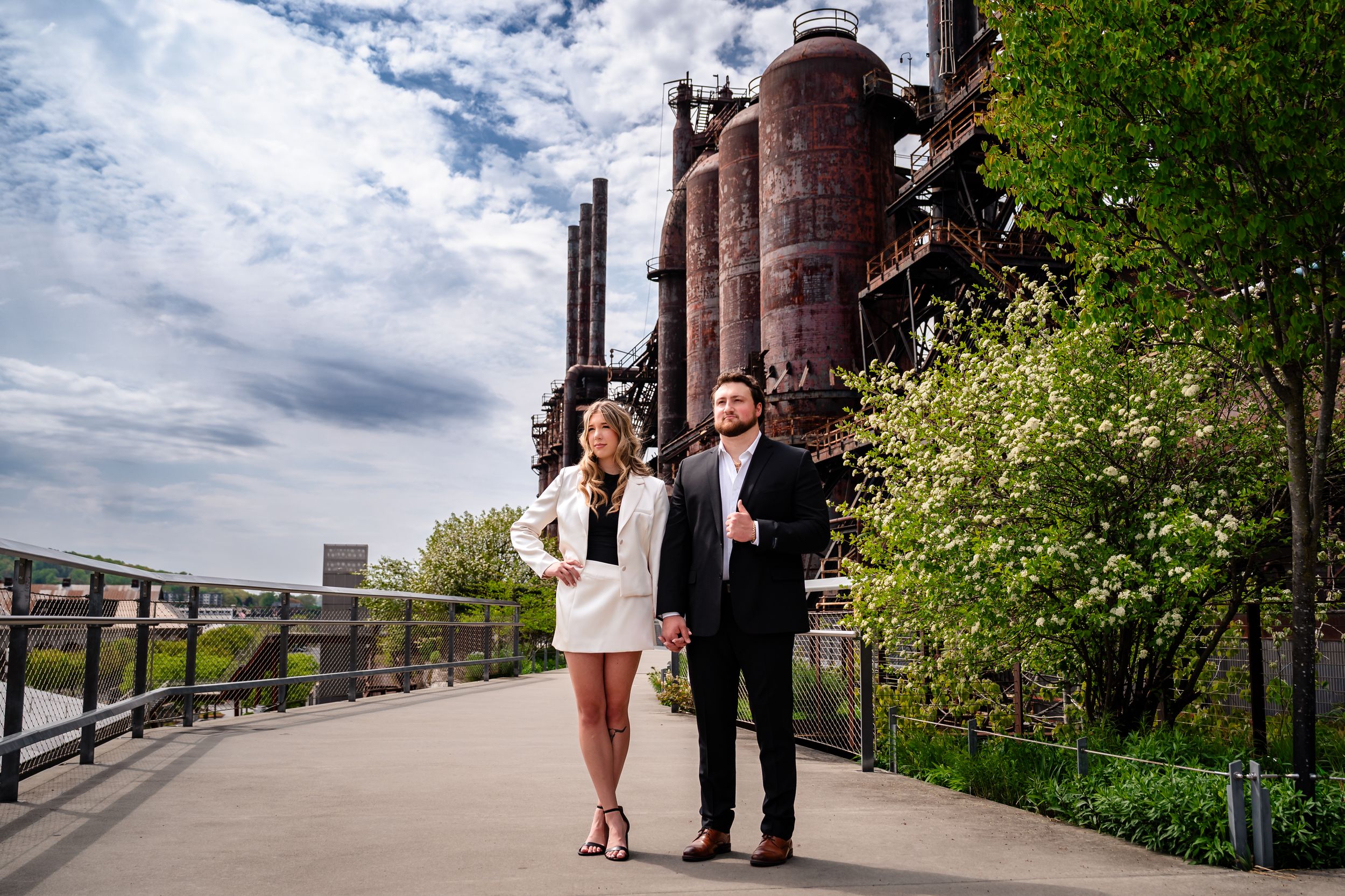 Elegant couple strolling on a walkway with towering rusty industrial blast furnaces behind them.