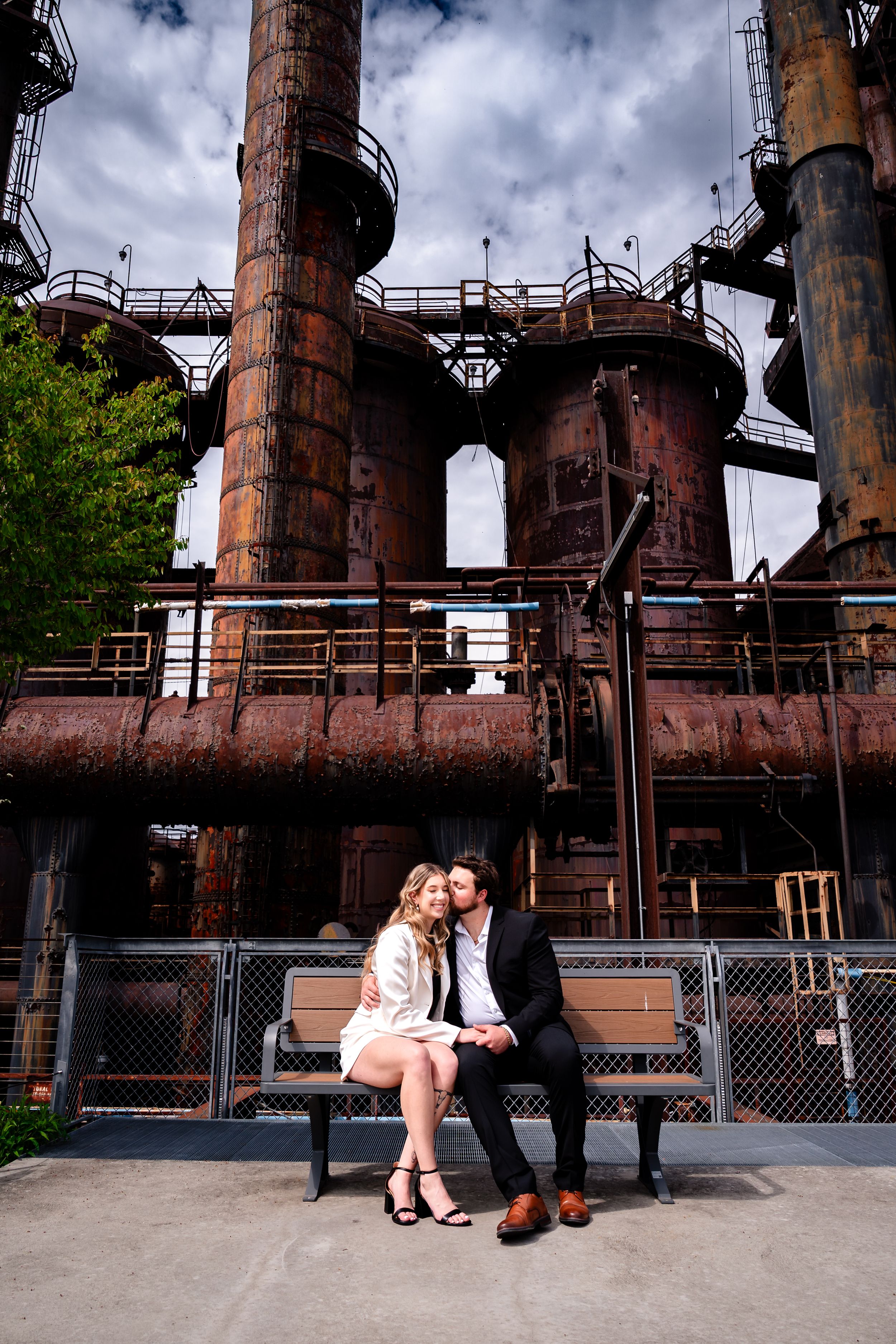 Couple sitting closely on a bench in front of massive rusty industrial blast furnace towers.