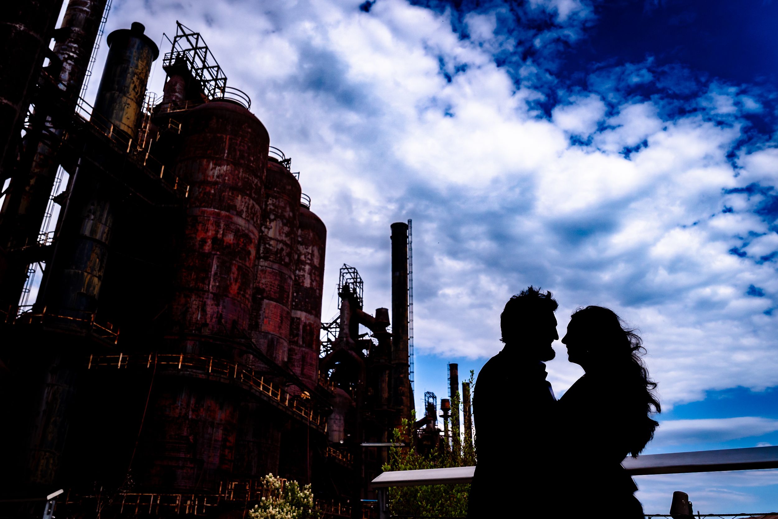 Silhouette of a romantic couple against dramatic cloudy sky with rustic industrial steel structures at dusk.