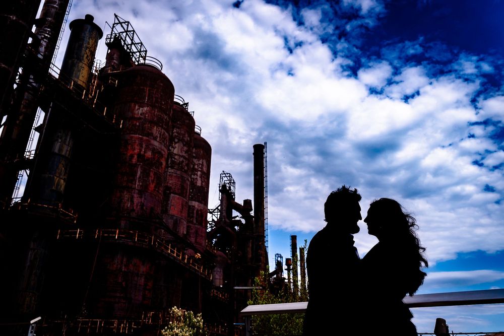 Silhouette of a romantic couple against dramatic cloudy sky with rustic industrial steel structures at dusk.