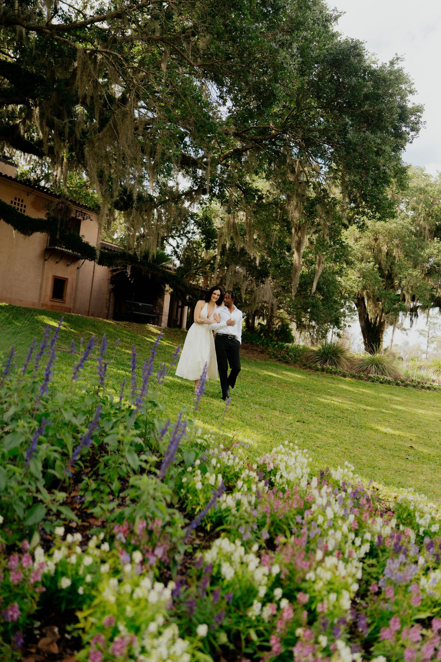 A bride and groom walk hand in hand through lush gardens with colorful flowers and moss-draped oak trees at an outdoor venue.