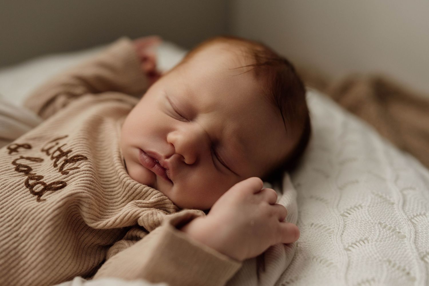 Sleeping newborn baby wrapped in sage green knit swaddle, cradled in adult hands on a wooden prop.