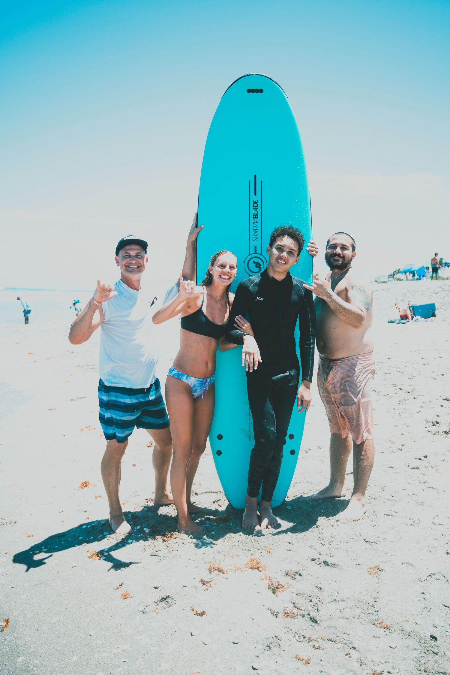 Four friends posing with a turquoise surfboard on a sunny beach, smiling and making hand gestures.