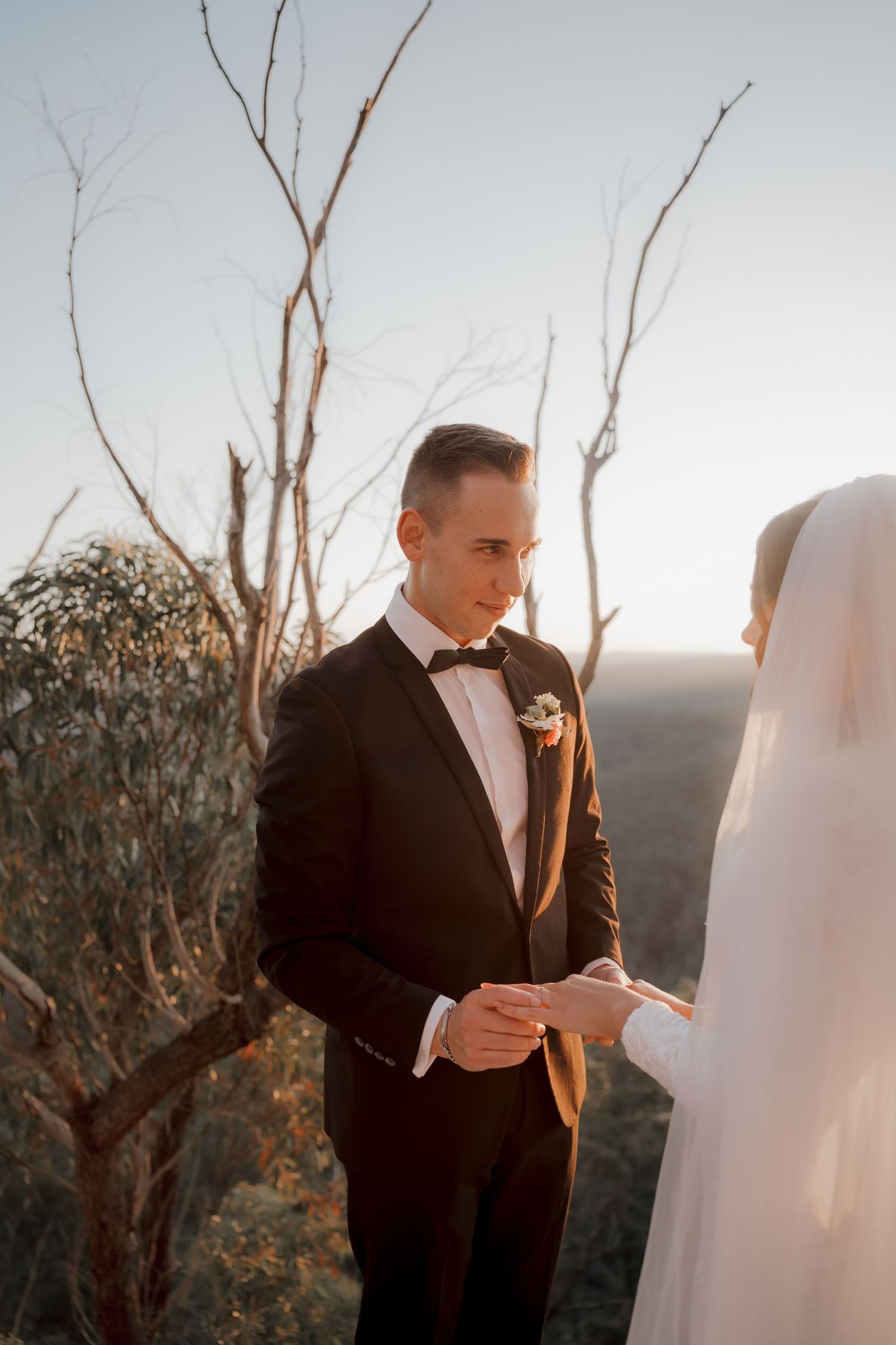 Groom in black tuxedo placing wedding ring on bride's finger during outdoor ceremony at sunset.