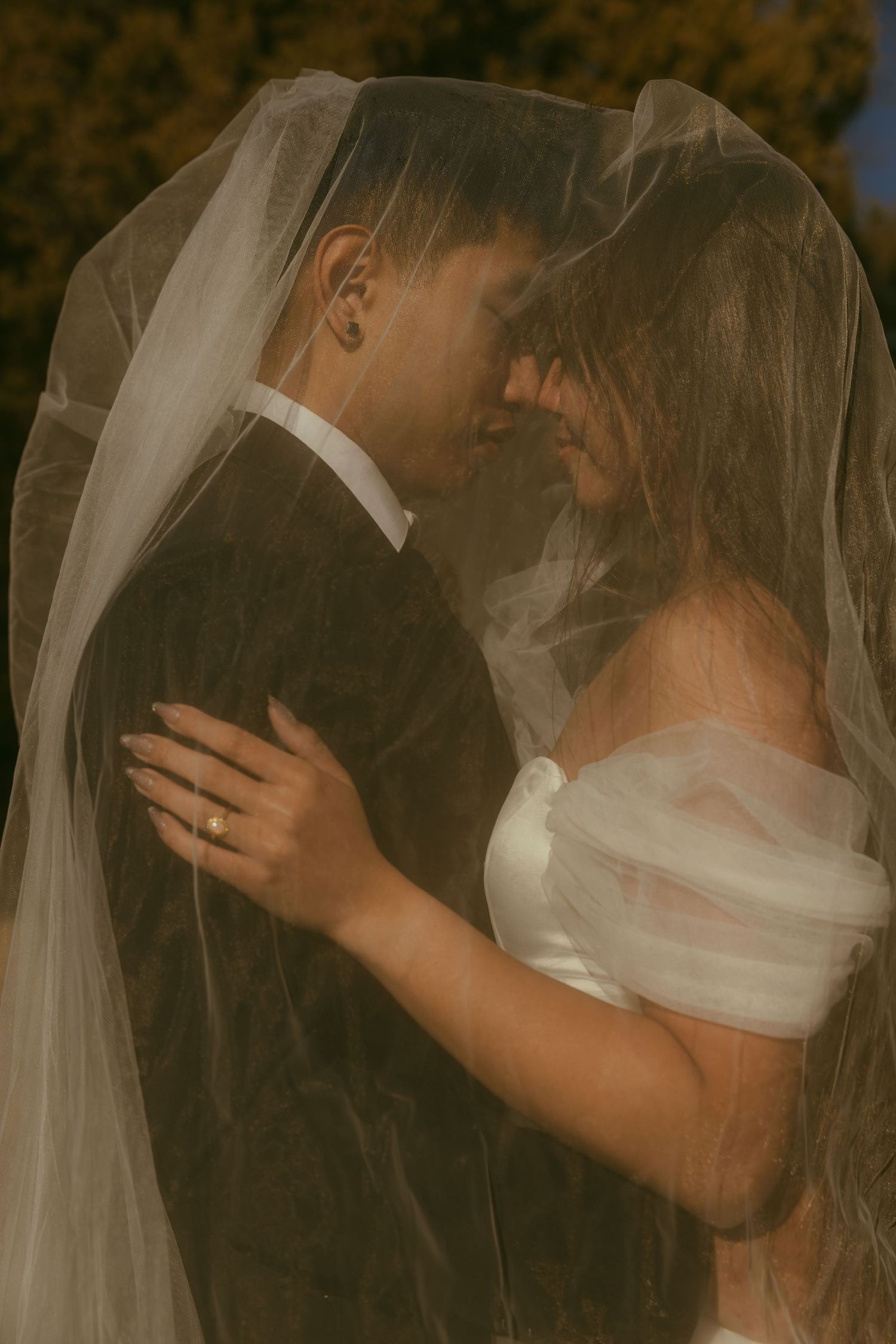 Bride and groom share an intimate moment under a flowing white veil, dressed in wedding attire with warm golden tones.