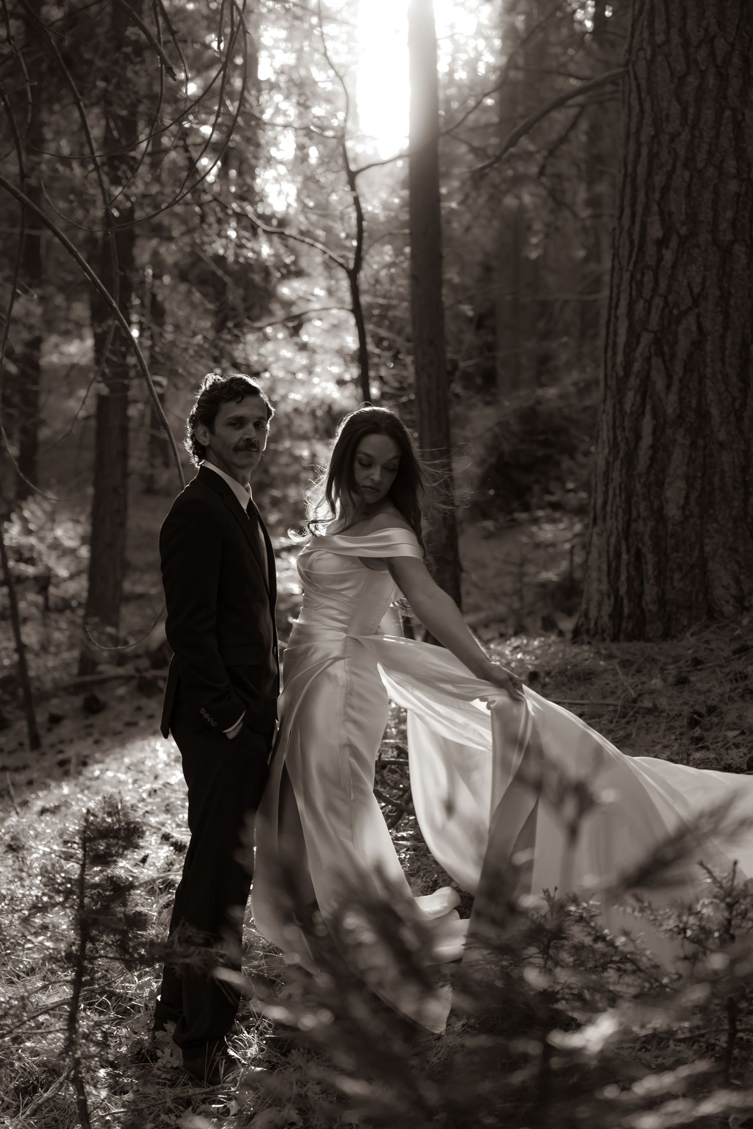 Bride in flowing white gown and groom in dark suit pose romantically in sunlit forest in sepia-toned wedding photo.